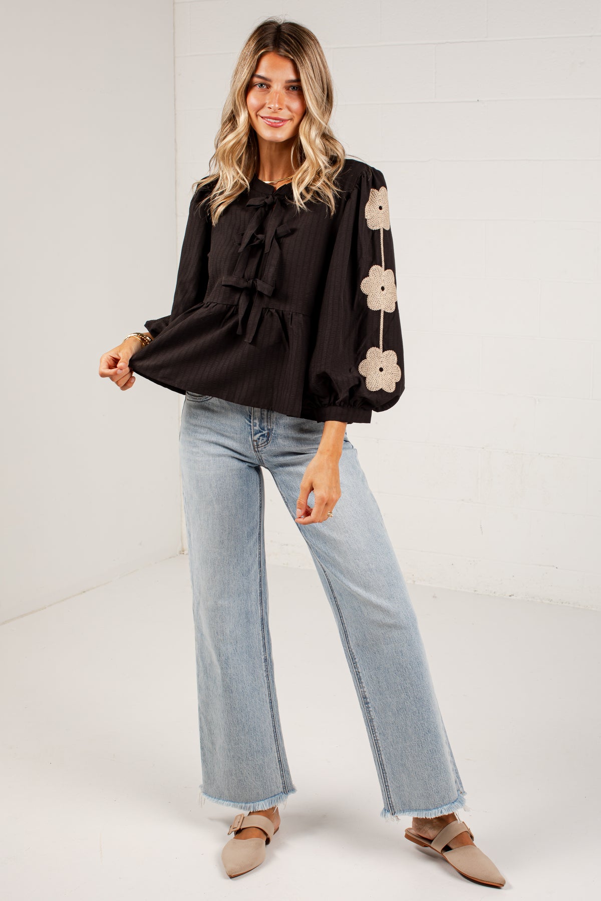A woman modeling a black long-sleeve blouse with a ruffled tie neckline and beige floral embroidery down one sleeve, paired with light-wash wide-leg jeans and tan flat shoes against a white studio background.