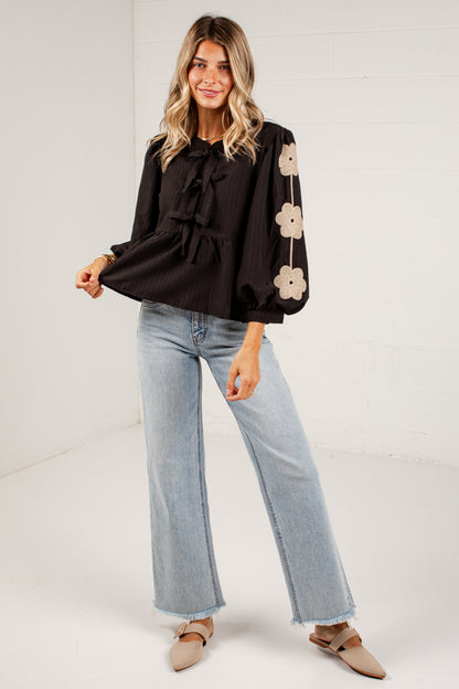 A woman modeling a black long-sleeve blouse with a ruffled tie neckline and beige floral embroidery down one sleeve, paired with light-wash wide-leg jeans and tan flat shoes against a white studio background.