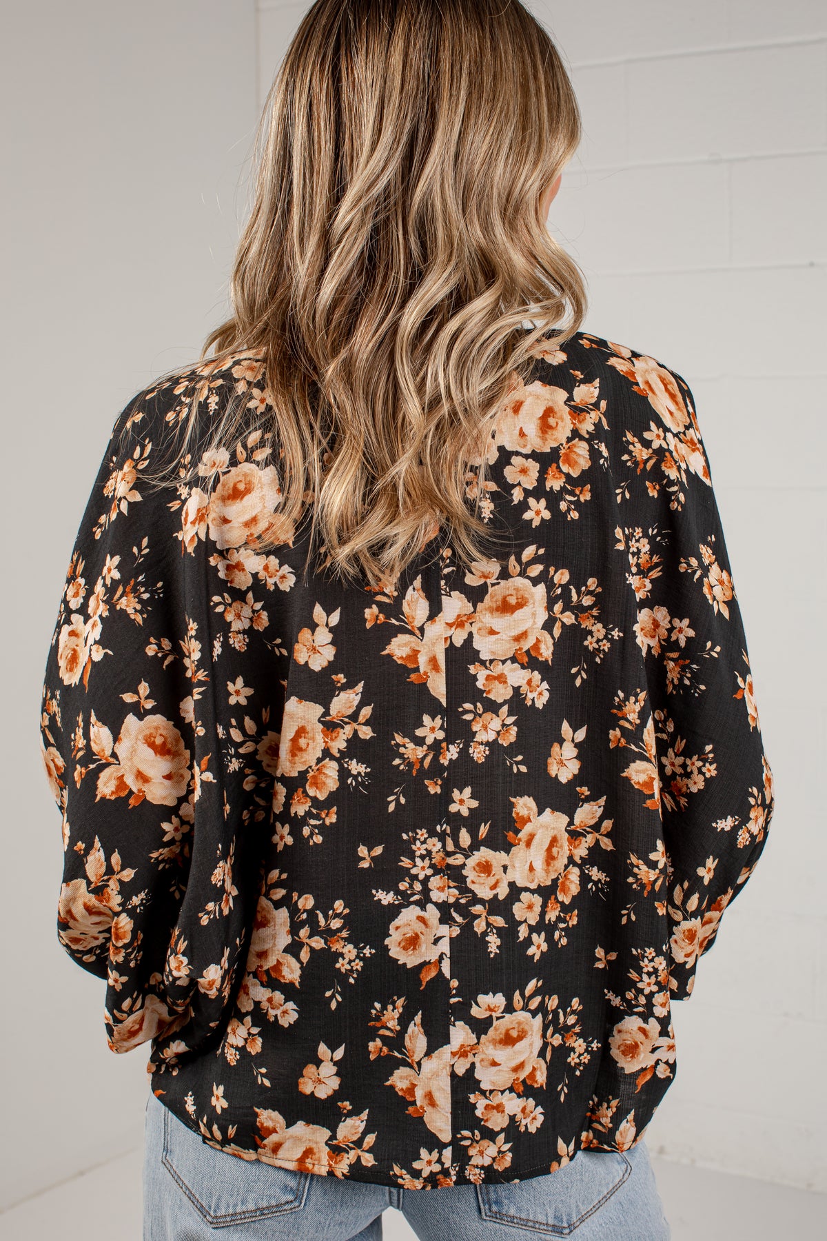 A woman viewed from behind wearing a black long-sleeve blouse with a warm-toned floral print in orange, beige, and cream, paired with light blue jeans against a white brick wall.