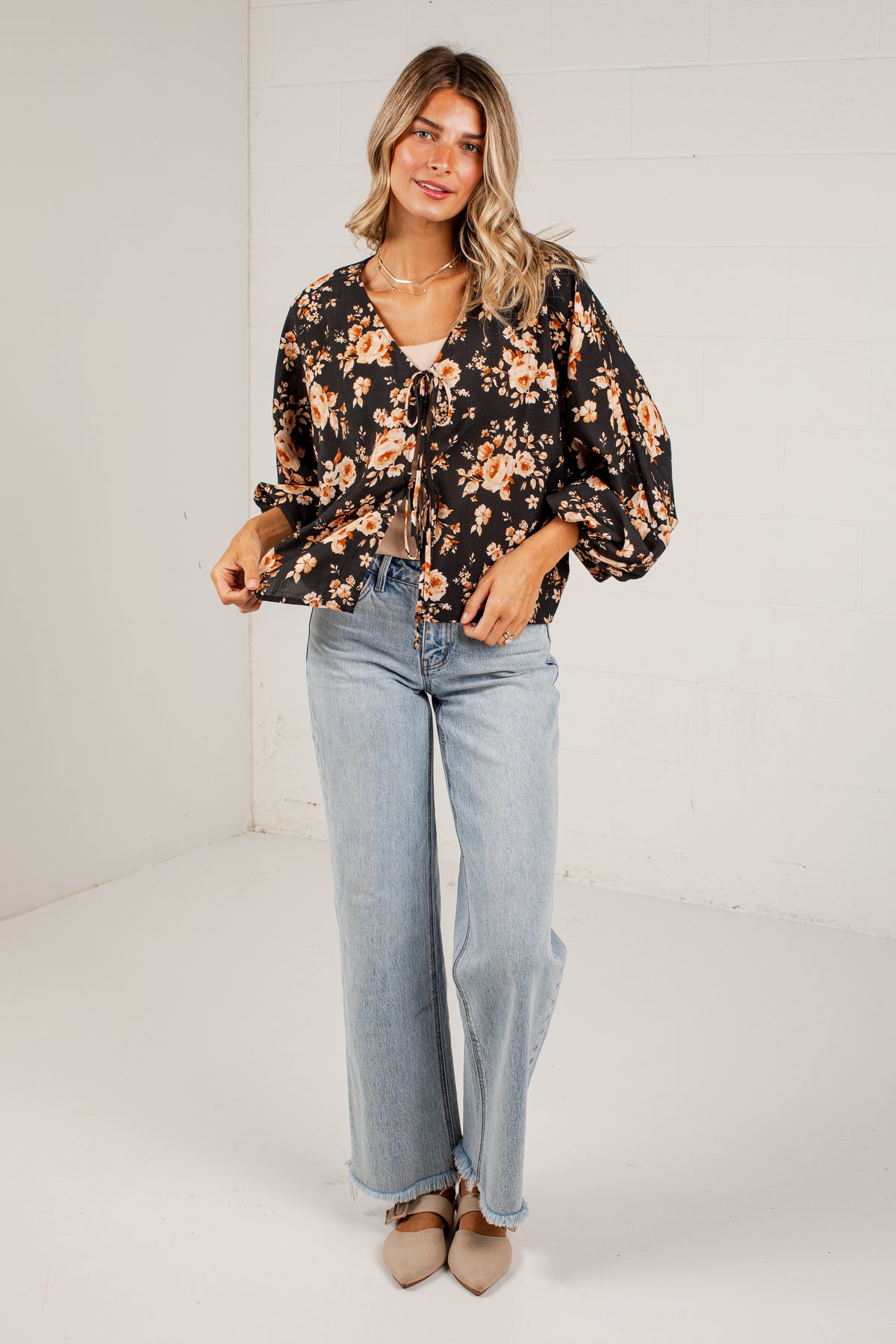A woman modeling a black long-sleeve blouse with a warm-toned floral print, paired with light-wash wide-leg jeans and beige pointed flats, standing against a white brick wall.