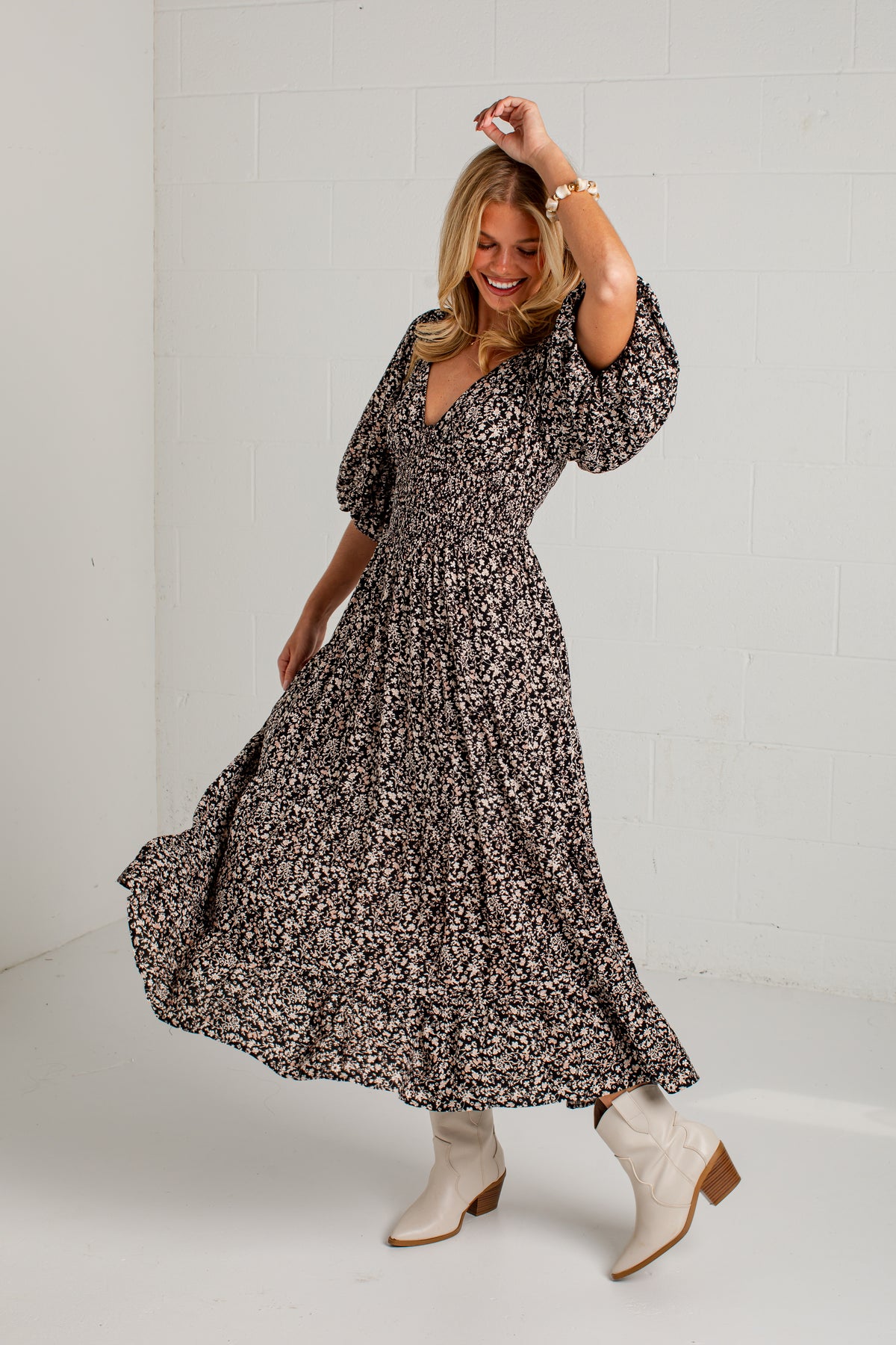 A woman modeling a long-sleeve, V-neck maxi dress with a dense black and white floral print, paired with white ankle boots, posing in a bright studio setting.