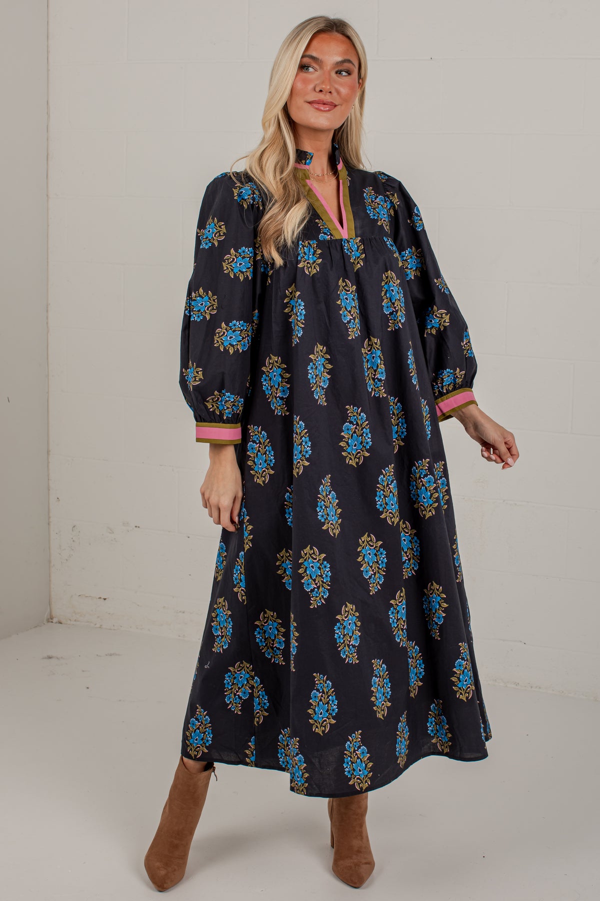 A woman modeling a long-sleeved black maxi dress with a blue and gold floral pattern and pink trim on the cuffs, paired with brown suede boots against a plain white studio background.