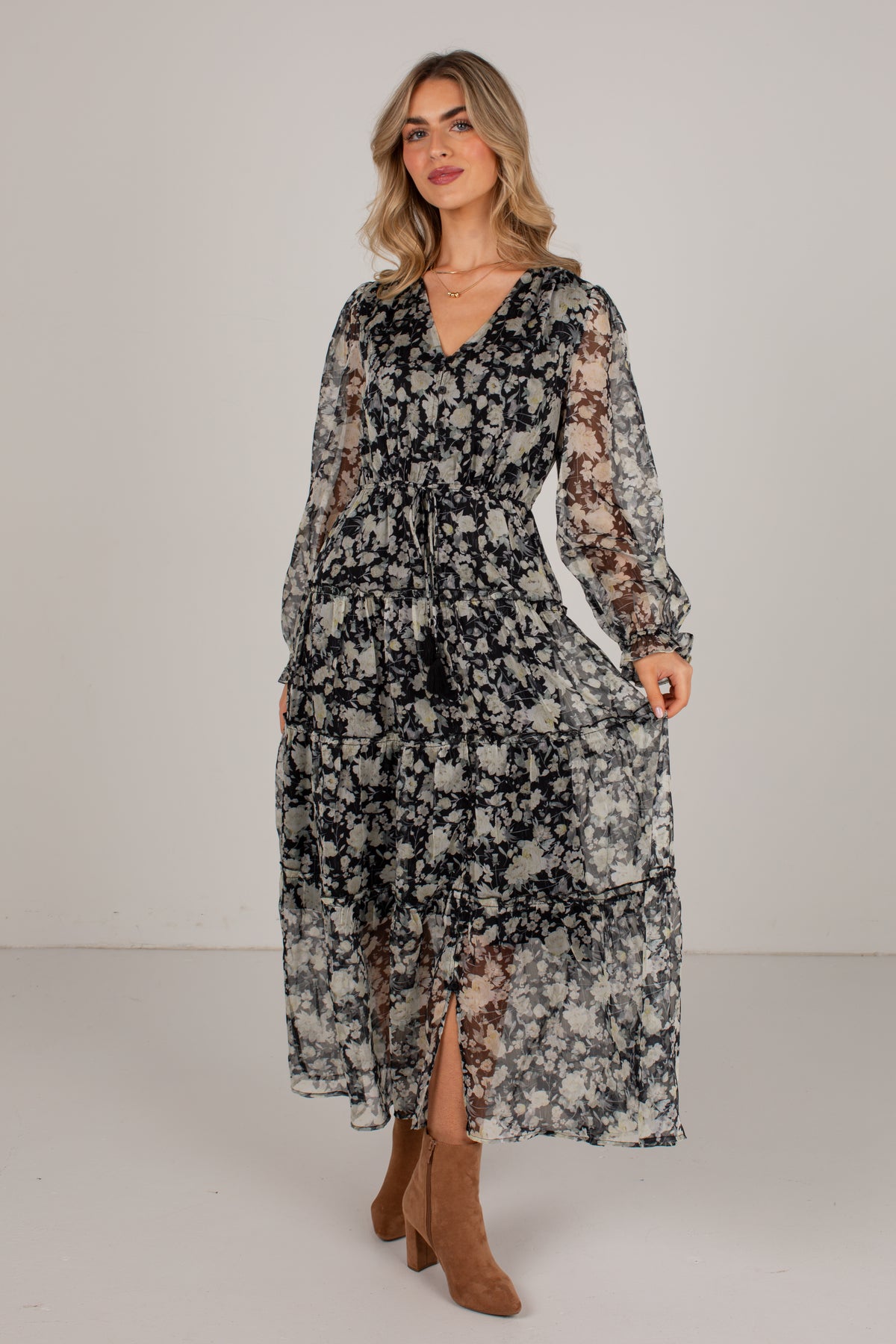A woman modeling a long-sleeve midi dress with a black and beige floral pattern, sheer lace sleeves, and a V-neckline, paired with brown suede boots against a plain studio background.