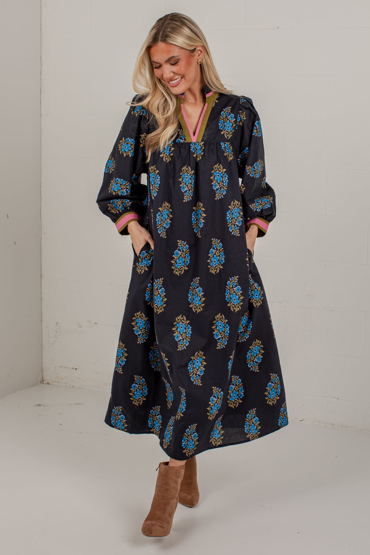 A woman modeling a long-sleeved black maxi dress with a blue and gold floral pattern, paired with brown suede boots, in a studio setting.