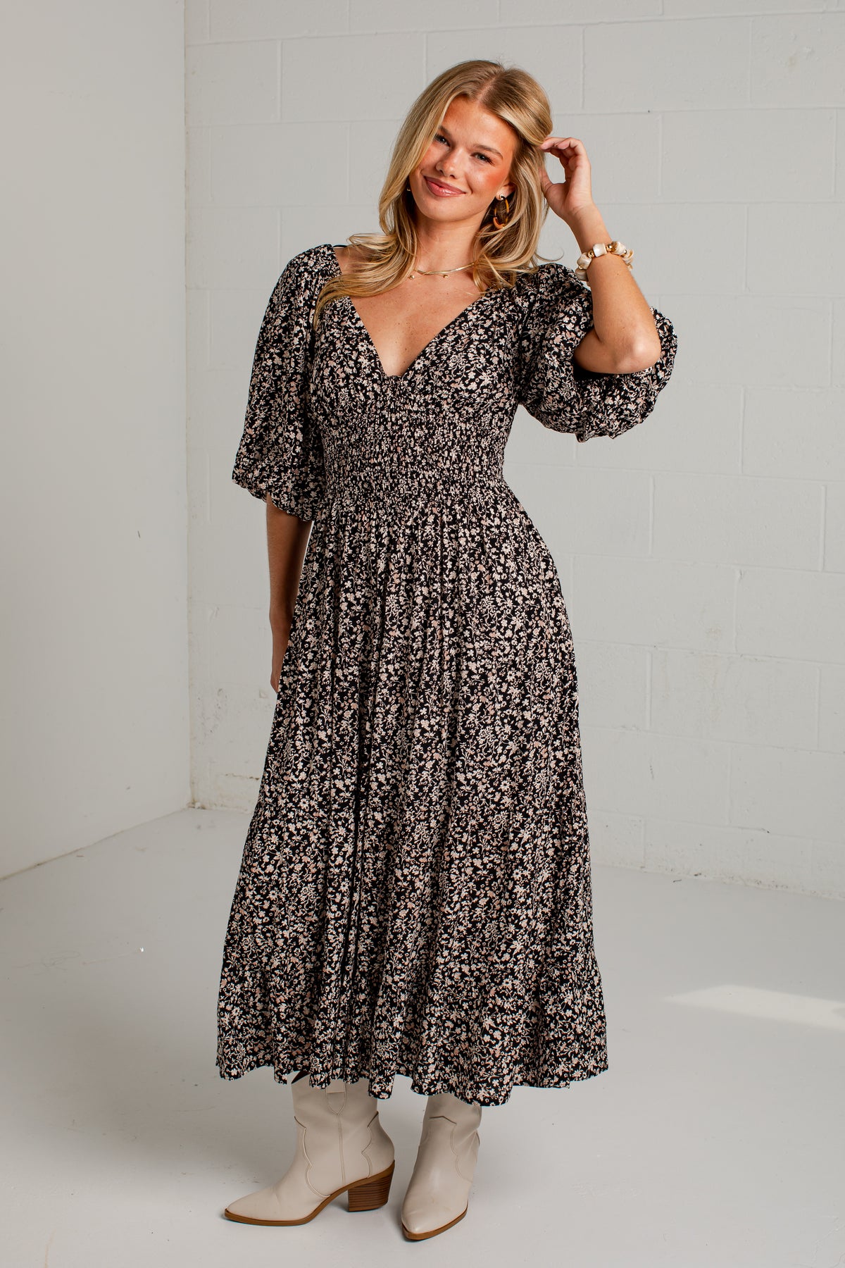 A woman modeling a black and beige floral print midi dress with puff sleeves and a V-neckline, paired with beige ankle boots in a bright studio setting.