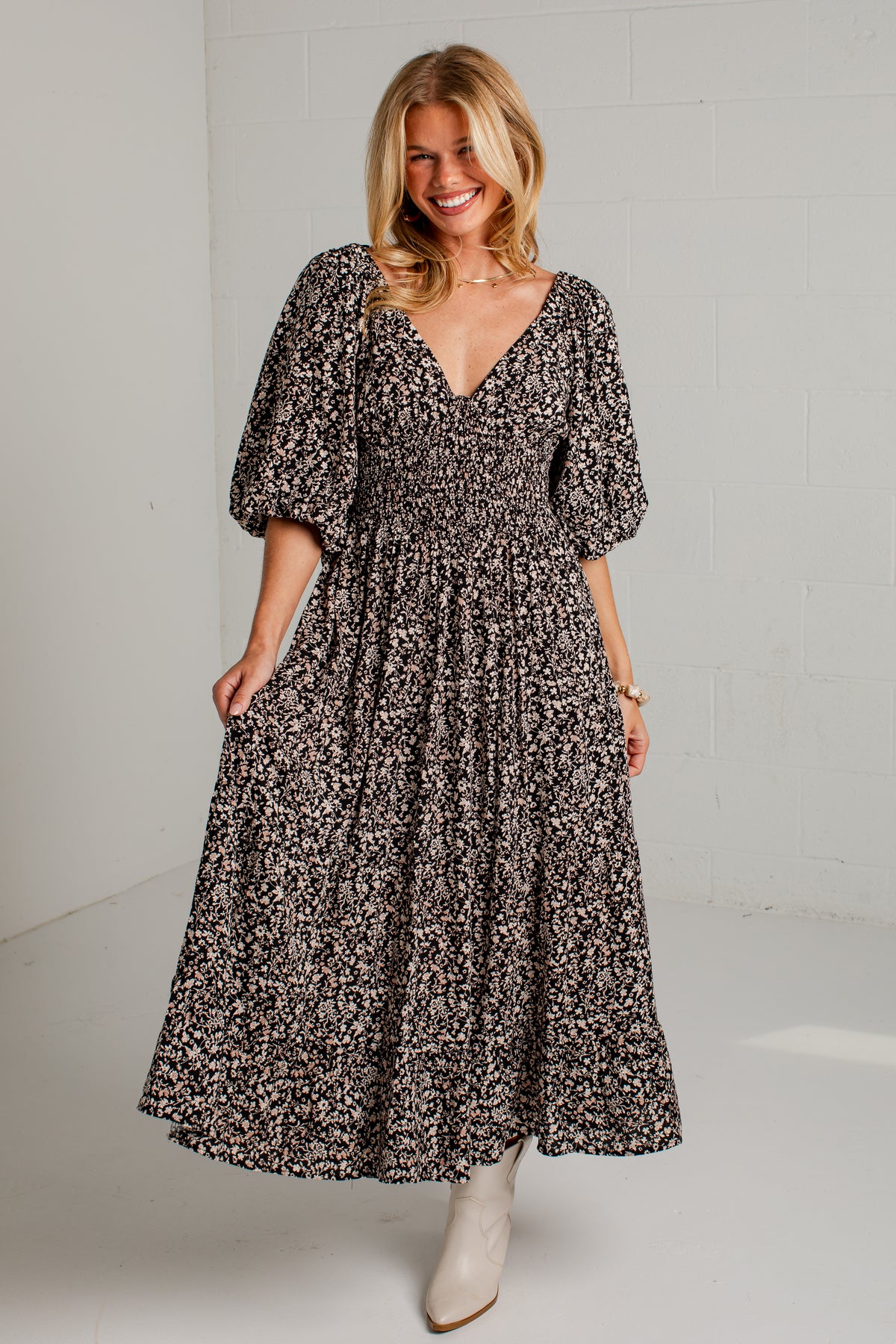 A woman modeling a black and white floral print maxi dress with puff sleeves and a smocked waist, paired with beige ankle boots, in a bright studio setting.