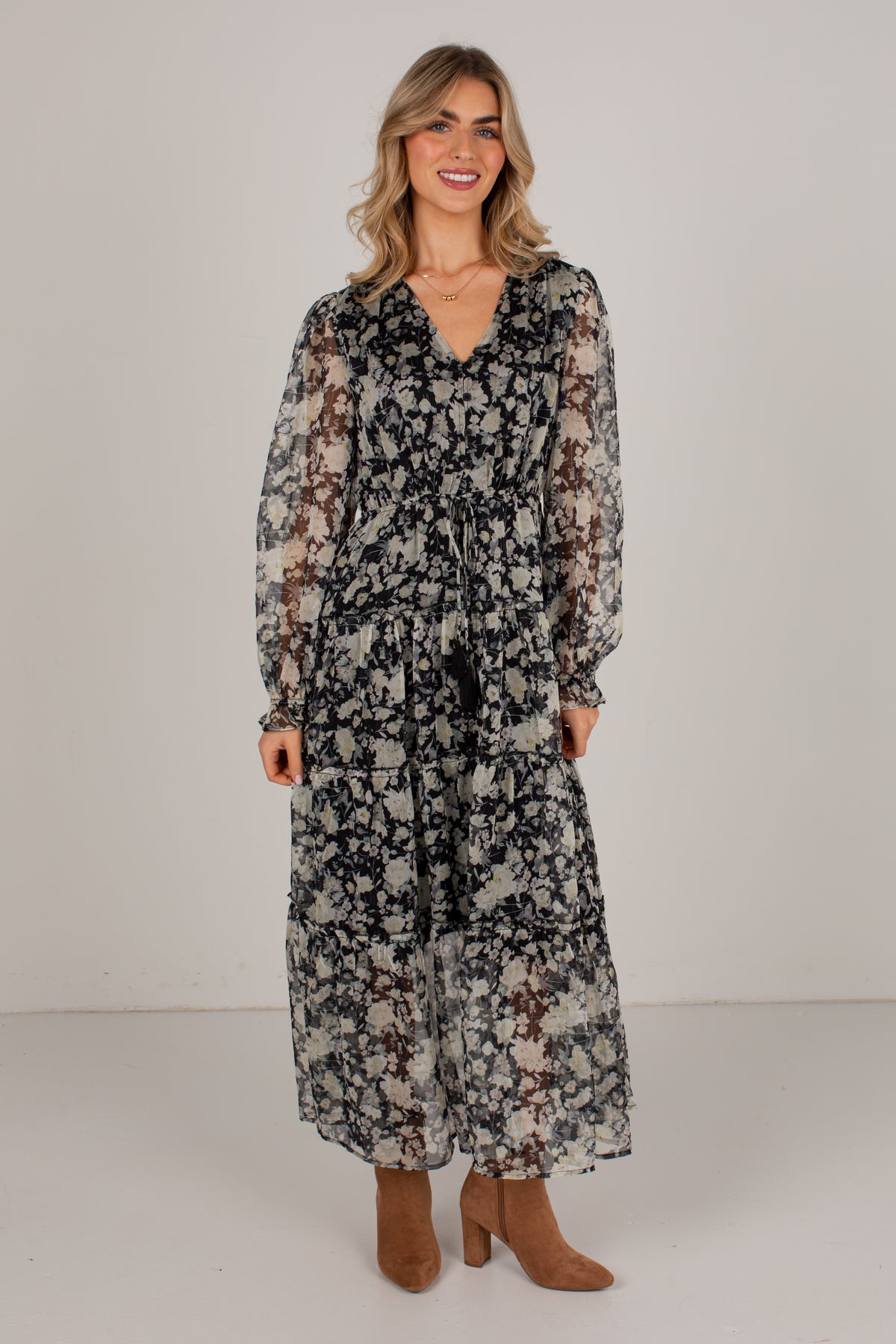 A woman modeling a long-sleeve midi dress with a black, gray, and brown floral pattern, paired with brown suede boots, against a plain light gray studio background.