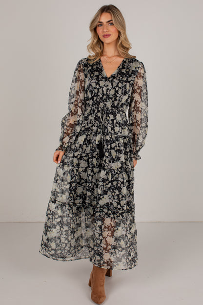 A woman modeling a long-sleeve midi dress with a black, gray, and beige floral pattern, standing against a plain light gray studio background.