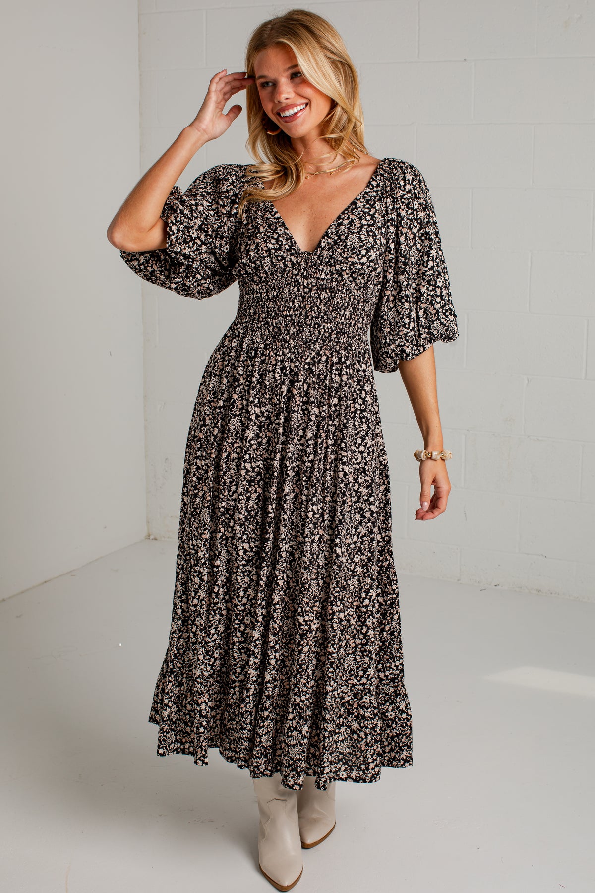 A woman modeling a black and beige floral print maxi dress with puff sleeves and a smocked bodice, paired with beige ankle boots in a bright studio setting.