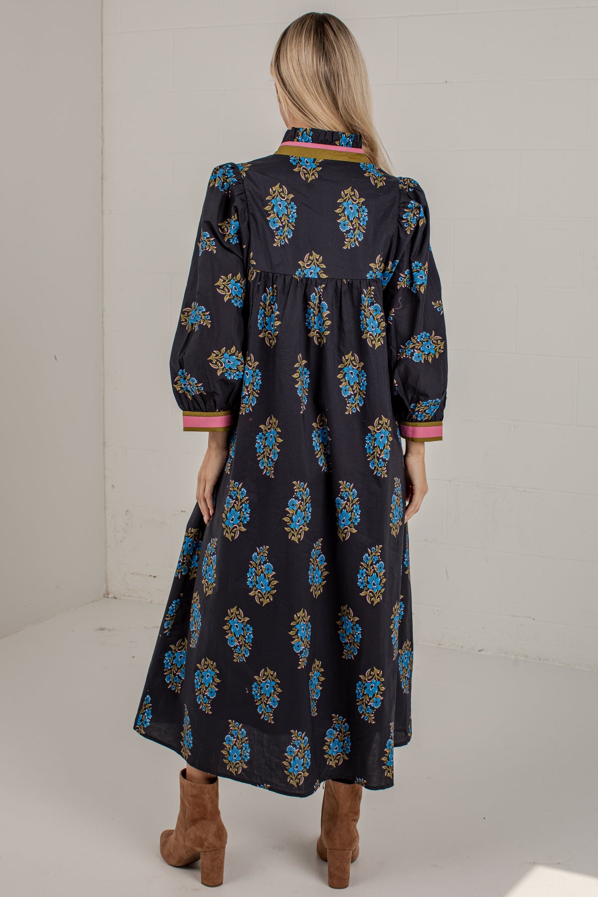 A woman viewed from behind wearing a long black midi dress with a colorful blue, gold, and pink floral pattern, paired with brown ankle boots in a white studio setting.