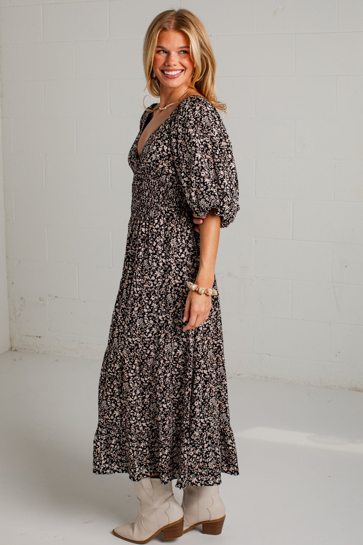 A woman modeling a long black and white floral print midi dress with puff sleeves, paired with beige ankle boots, standing against a white brick wall.