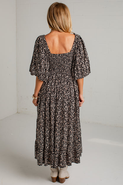 A woman viewed from behind wearing a black and beige floral maxi dress with puff sleeves and a smocked bodice, standing in a bright white room.