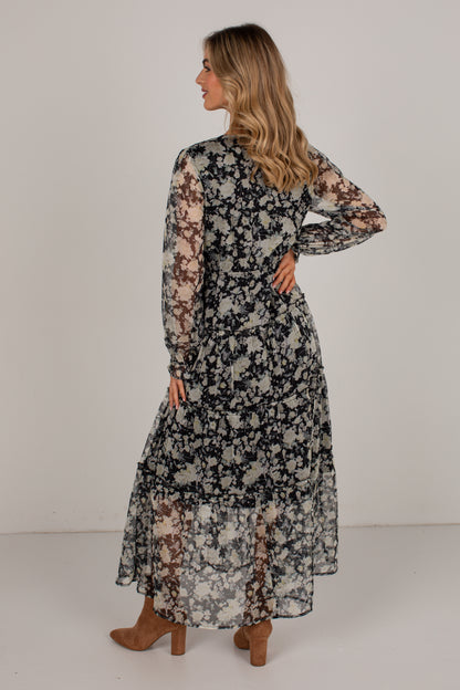 A woman modeling a long-sleeve midi dress with a black, white, and brown floral pattern, shown from a back-three-quarter view against a plain light gray studio background.