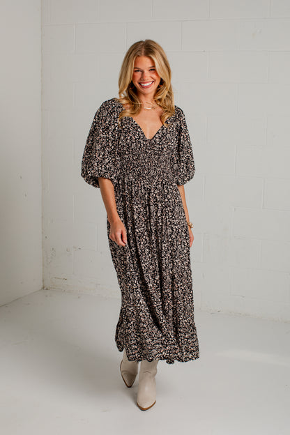 A woman modeling a black and beige floral print maxi dress with short puff sleeves and a V-neckline, paired with beige ankle boots, standing against a white brick wall.