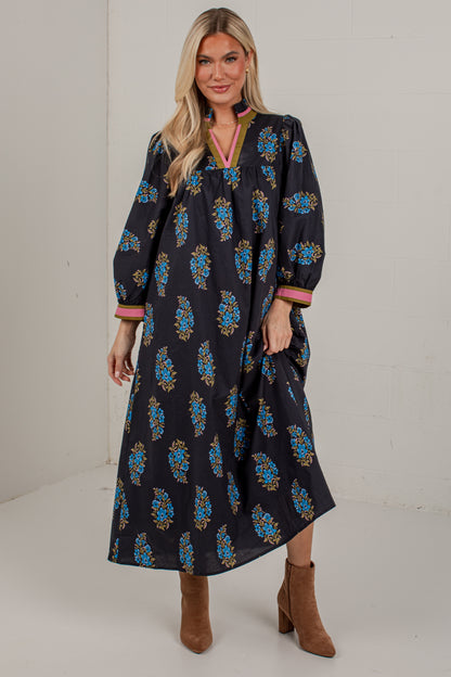 A woman modeling a long-sleeved navy blue maxi dress with a blue, gold, and pink floral pattern, paired with brown ankle boots, set against a plain off-white studio background.