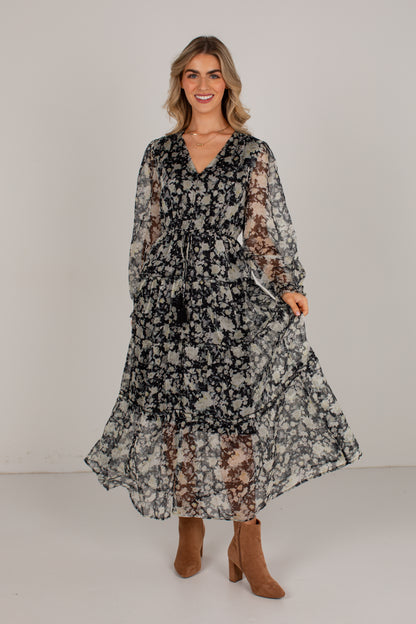 A woman modeling a long-sleeved, tiered maxi dress with a black and white floral pattern, paired with brown suede ankle boots, against a plain light gray studio background.