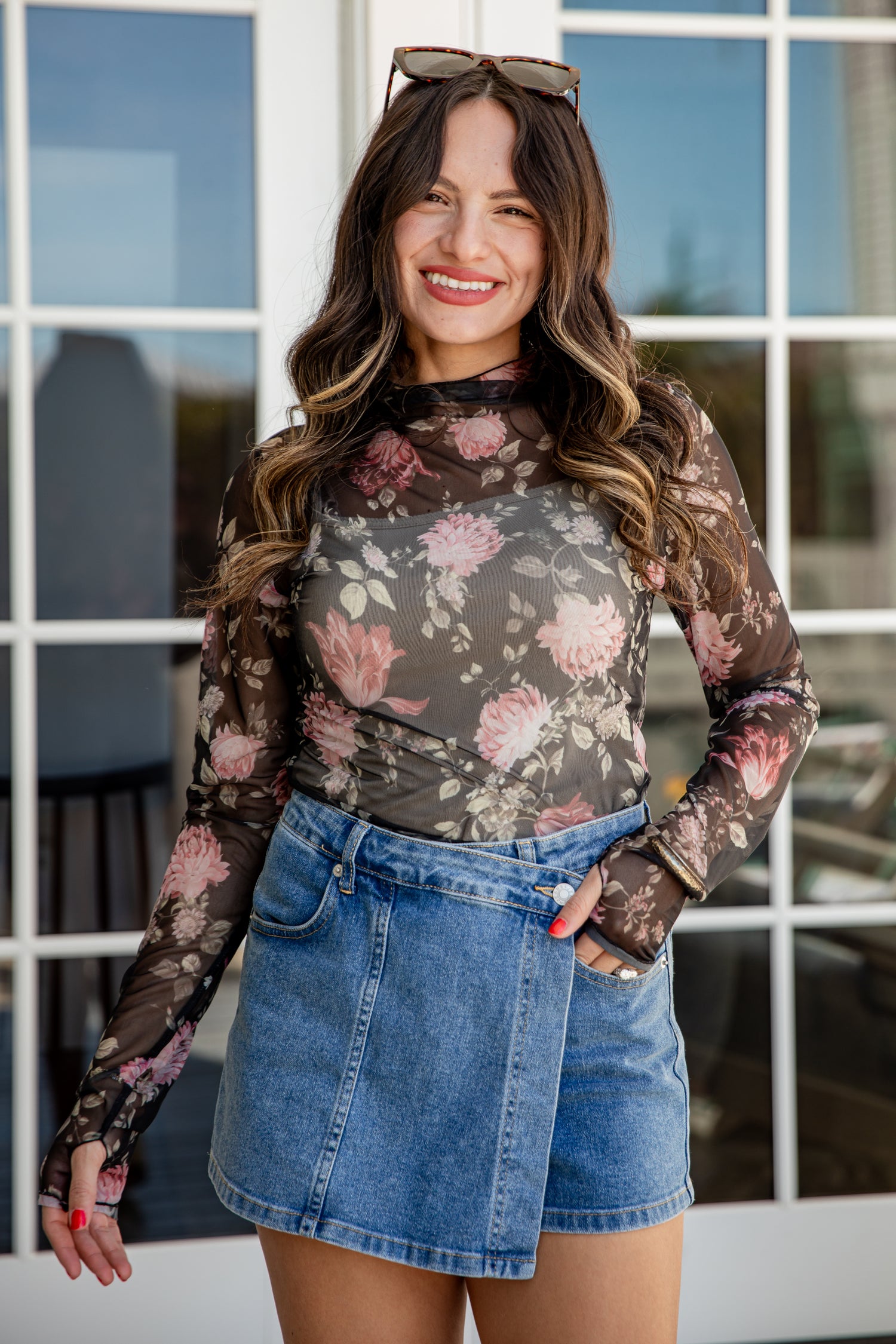 Woman wearing a floral long-sleeve top and denim skirt standing in front of a window.