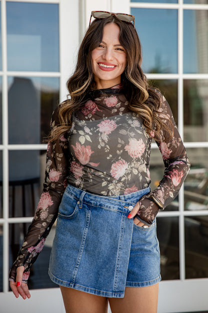 Woman wearing a floral long-sleeve top and denim skirt standing in front of a window.