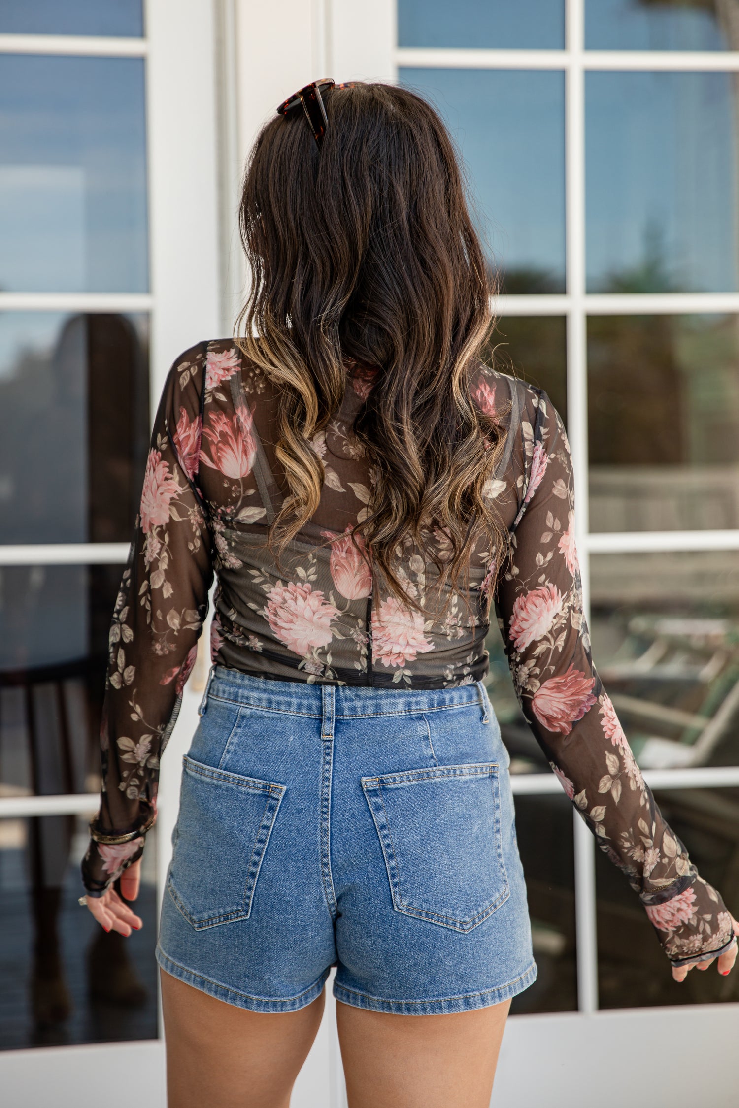Woman wearing a floral long-sleeve top and denim shorts standing in front of a glass door.