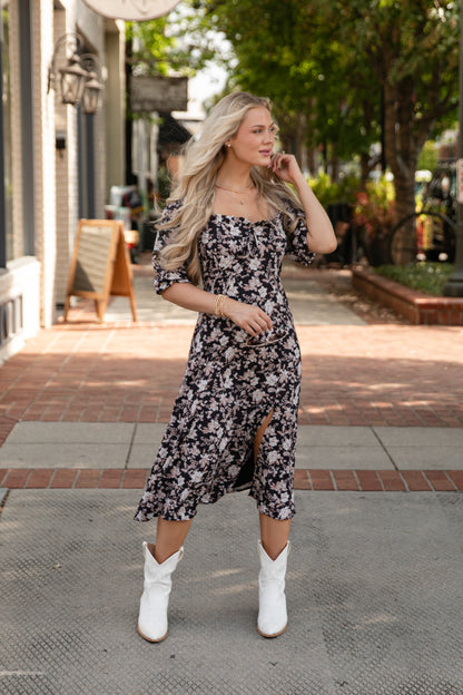 Woman in a floral dress and white boots standing on a sidewalk.