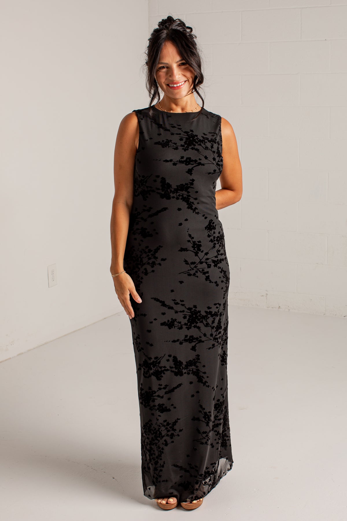 A woman modeling a sleeveless black maxi dress with a tonal floral lace overlay, standing against a white studio wall.