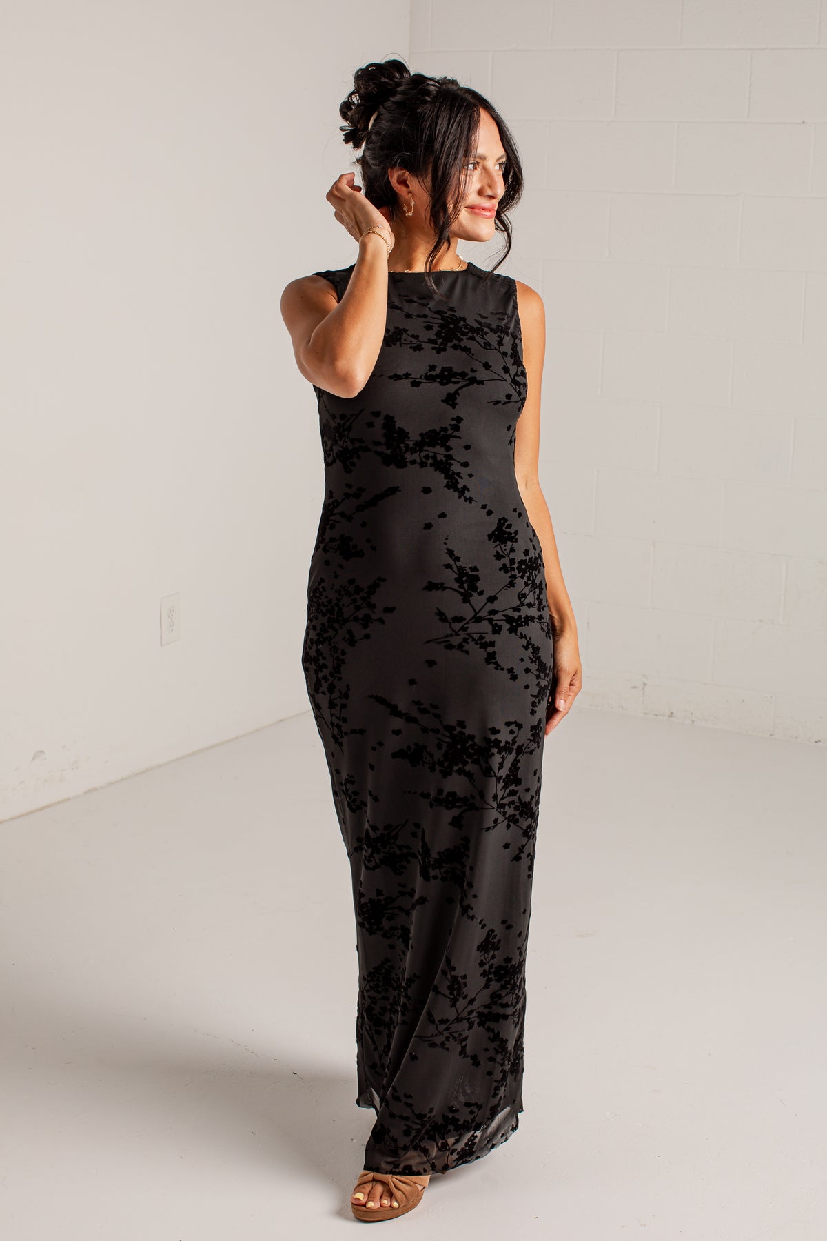 A woman modeling a sleeveless black maxi dress with a tonal floral lace overlay, standing in a bright white studio.