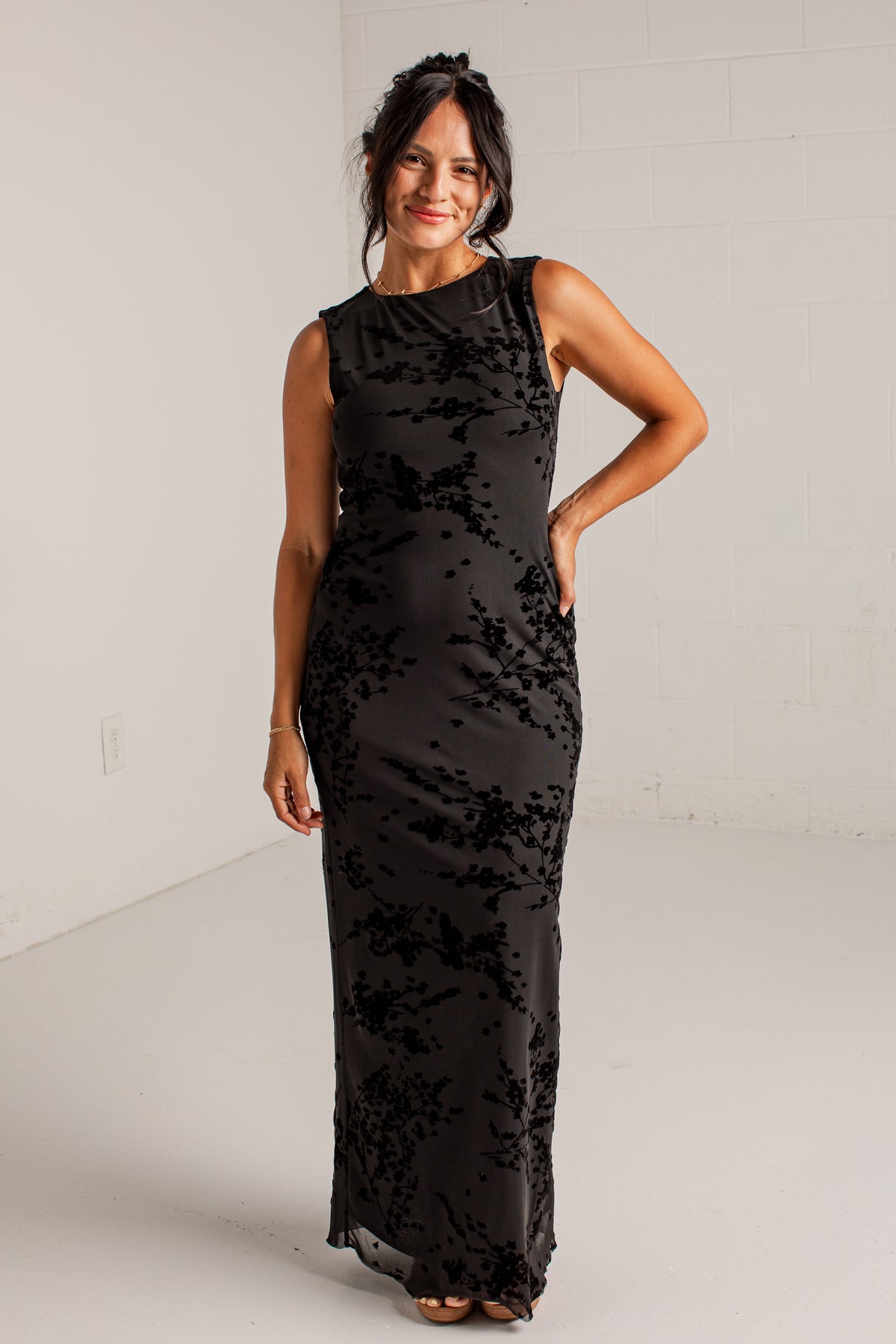 A woman modeling a sleeveless black maxi dress with a textured, slightly shiny floral or abstract pattern, standing in a bright white studio.