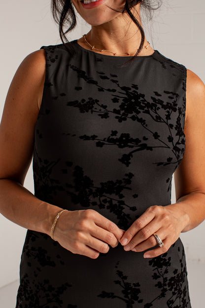 A sleeveless black mesh dress with a black floral branch pattern, worn by a model. The dress features a gathered neckline and a textured, ribbed bodice and skirt.