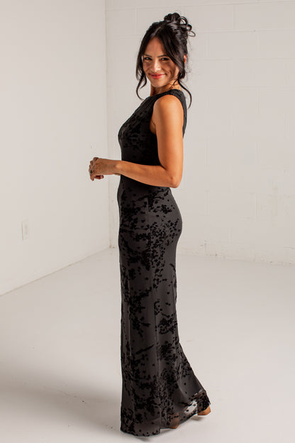 A woman modeling a sleeveless black maxi dress with a textured, tonal floral pattern, standing in a minimalist white studio setting.