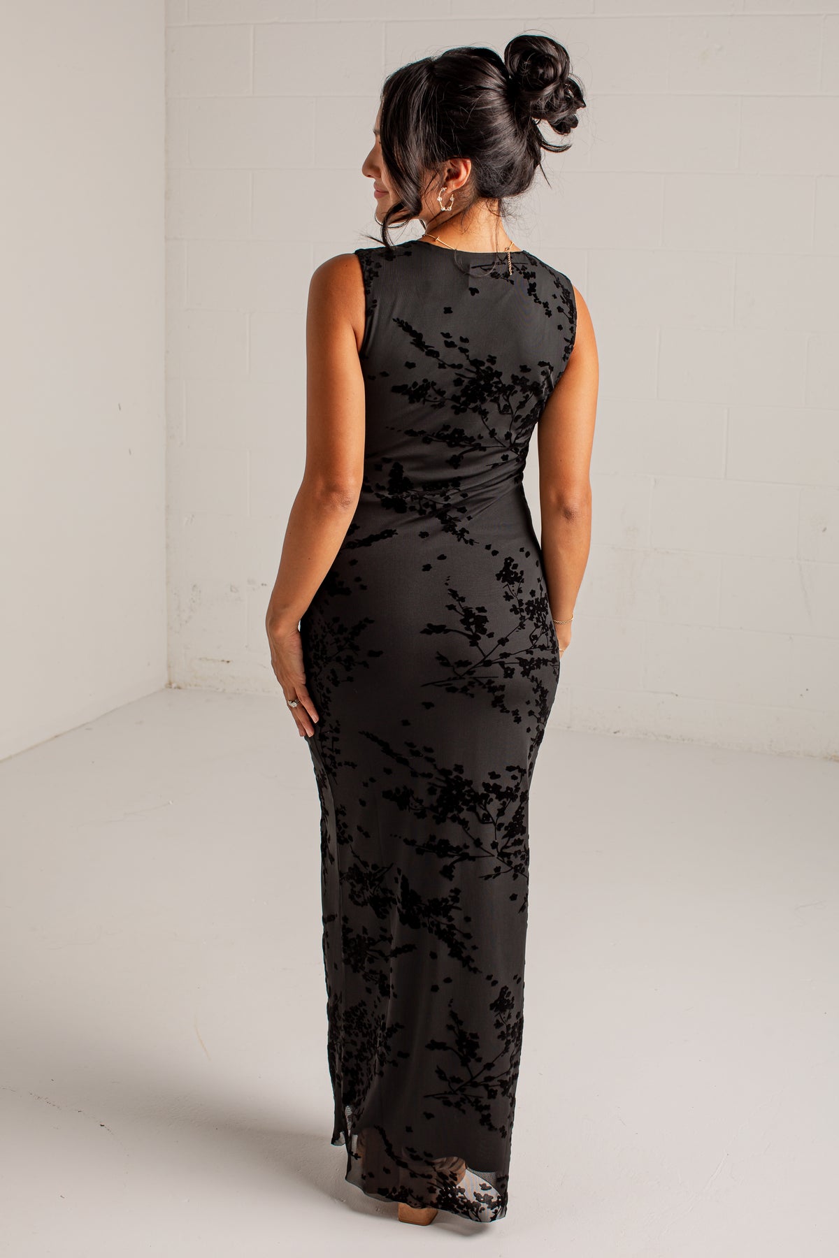A woman viewed from behind wearing a sleeveless black maxi dress with a tonal floral lace overlay, standing in a white studio corner.