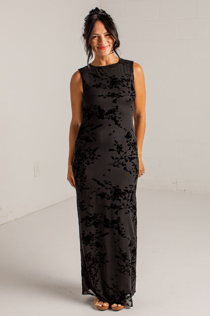 A woman modeling a sleeveless black maxi dress with a textured, tonal floral or abstract pattern, standing against a plain white studio wall.