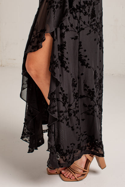 A close-up of a person wearing a black sheer floral maxi dress with ruffled layers and tan strappy heeled sandals, standing against a light neutral wall.