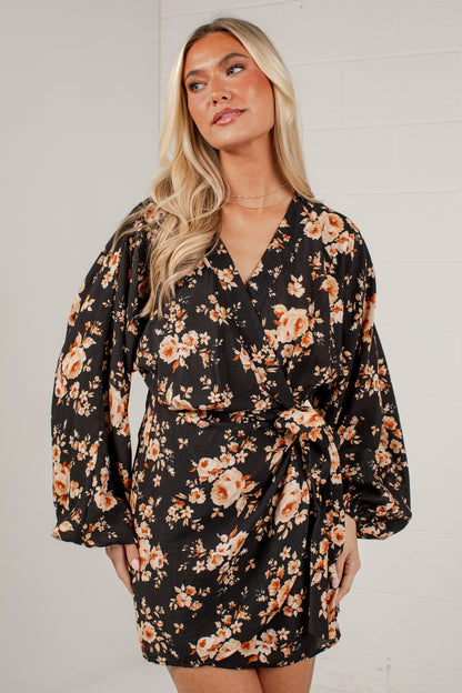 A woman wearing a black wrap-style mini dress with a warm-toned floral print in orange, peach, and beige, photographed against a light gray wall.