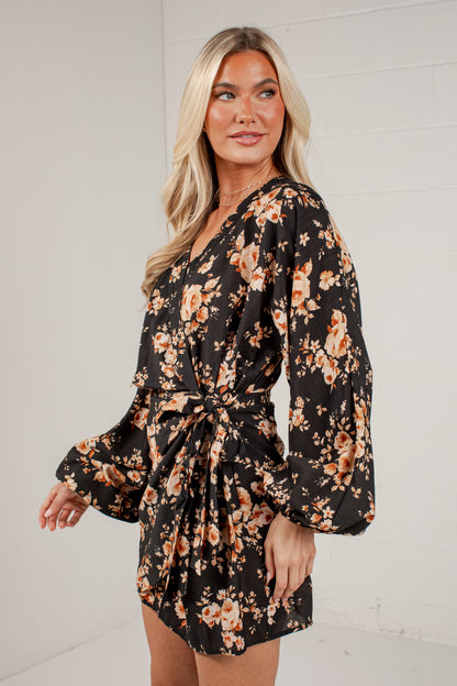 A woman modeling a black long-sleeve mini wrap dress with a warm orange and beige floral pattern, styled with a tie waist belt.