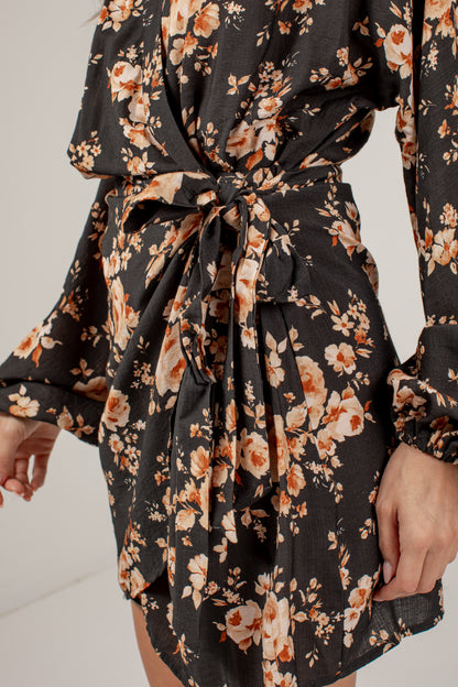 A close-up of a model wearing a black long-sleeve mini dress with a warm-toned floral print in orange, beige, and cream, featuring a tie waist belt.