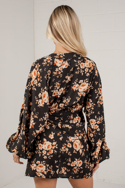A woman viewed from the back wearing a black long-sleeve mini dress with a warm-toned floral print in orange, peach, and beige, set against a white brick wall.