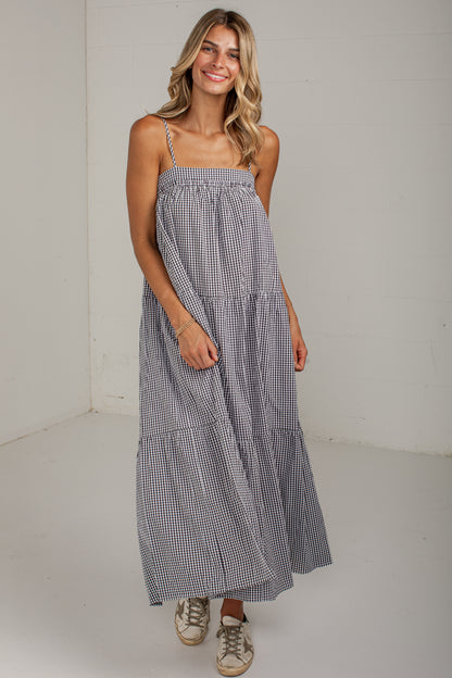 A woman modeling a sleeveless, tiered maxi dress with a small blue and white gingham pattern, standing against a plain off-white wall.