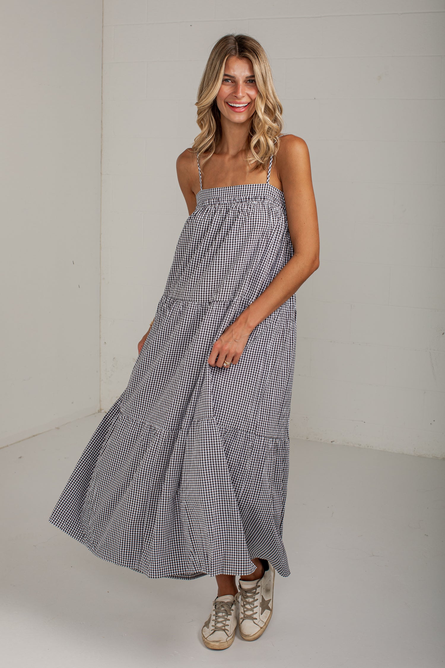 A woman modeling a sleeveless, tiered maxi dress with a small black and white gingham pattern, paired with beige sneakers, in a bright white studio setting.