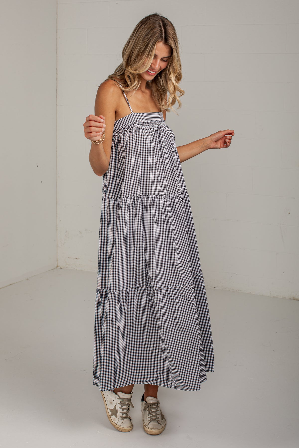 A woman wearing a sleeveless, tiered maxi dress with a small black and white gingham pattern, paired with beige sneakers, standing in a plain white room.