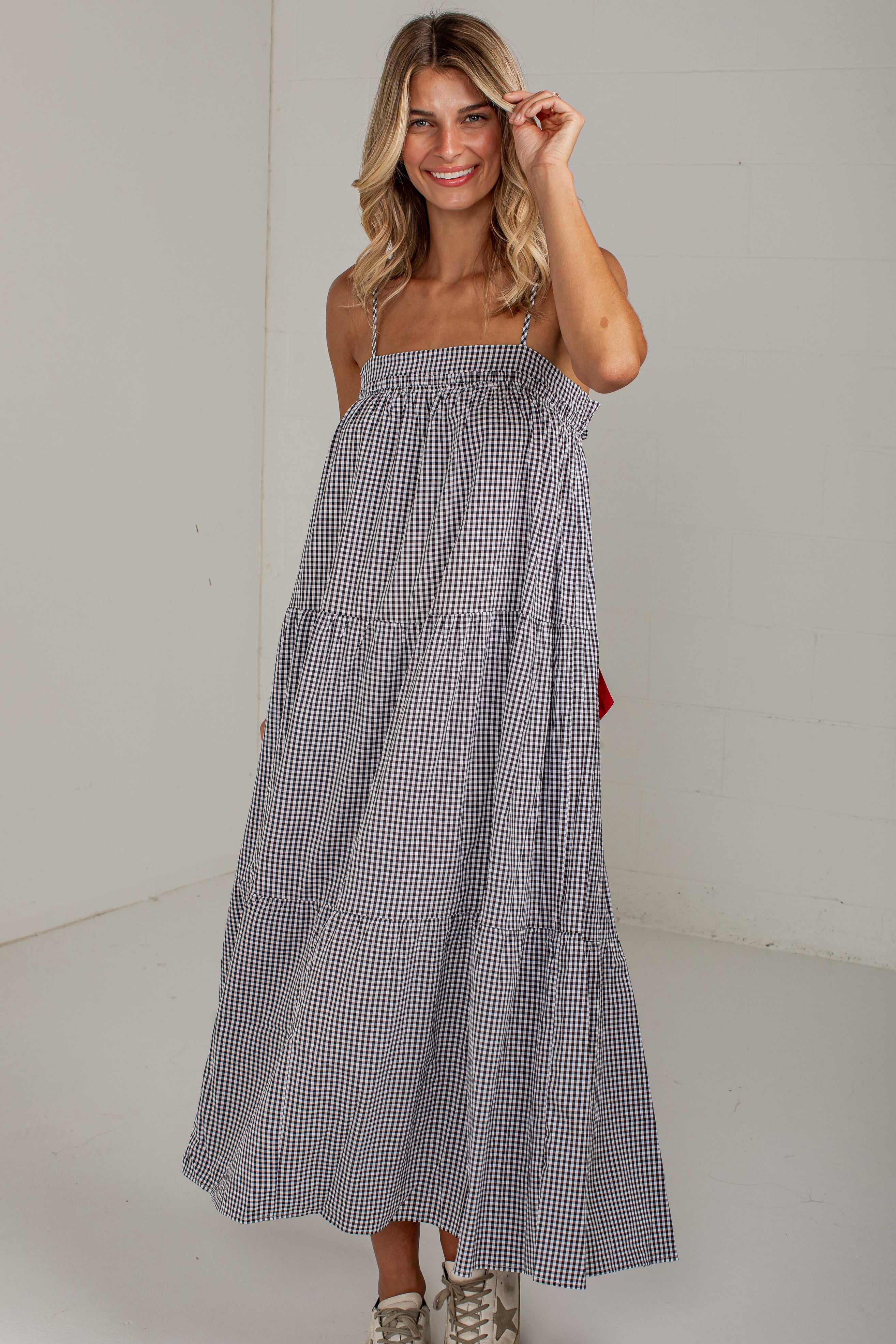 A woman modeling a sleeveless, tiered maxi dress with a small black and white gingham pattern, paired with beige sneakers, in a bright studio setting.