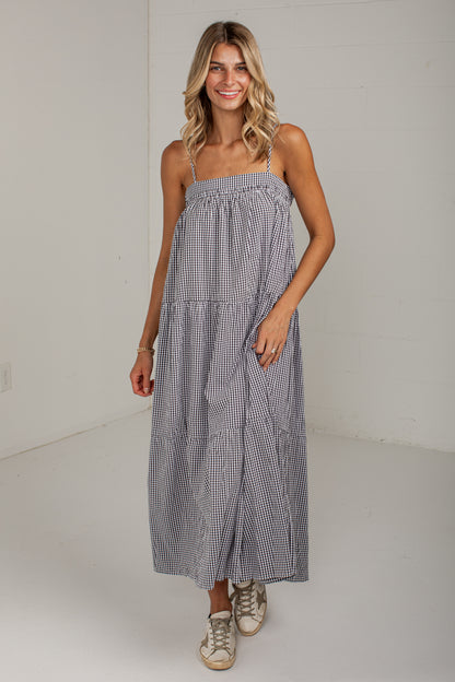 A woman modeling a sleeveless tiered maxi dress with a small blue and white gingham pattern, standing against a plain white studio wall.