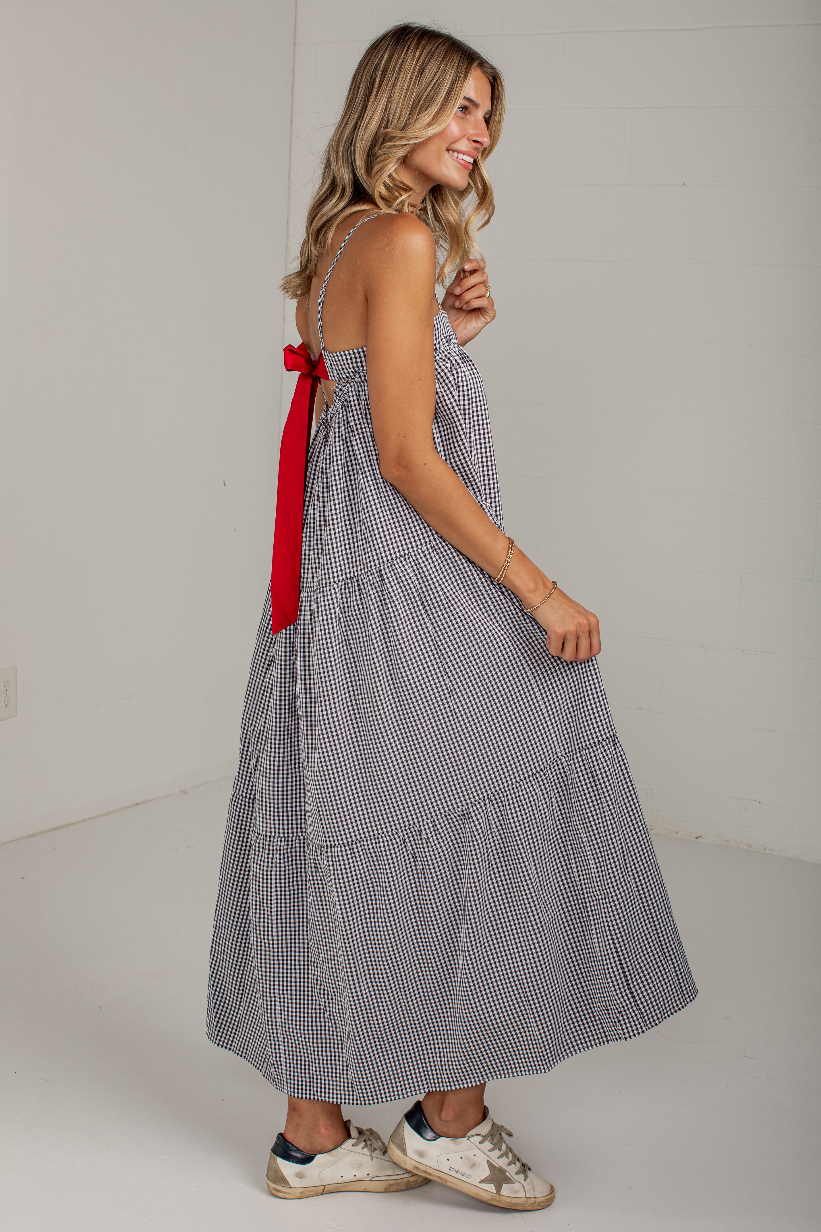 A woman modeling a sleeveless tiered maxi dress with a small blue and white gingham pattern and a contrasting red bow at the back, paired with white sneakers.