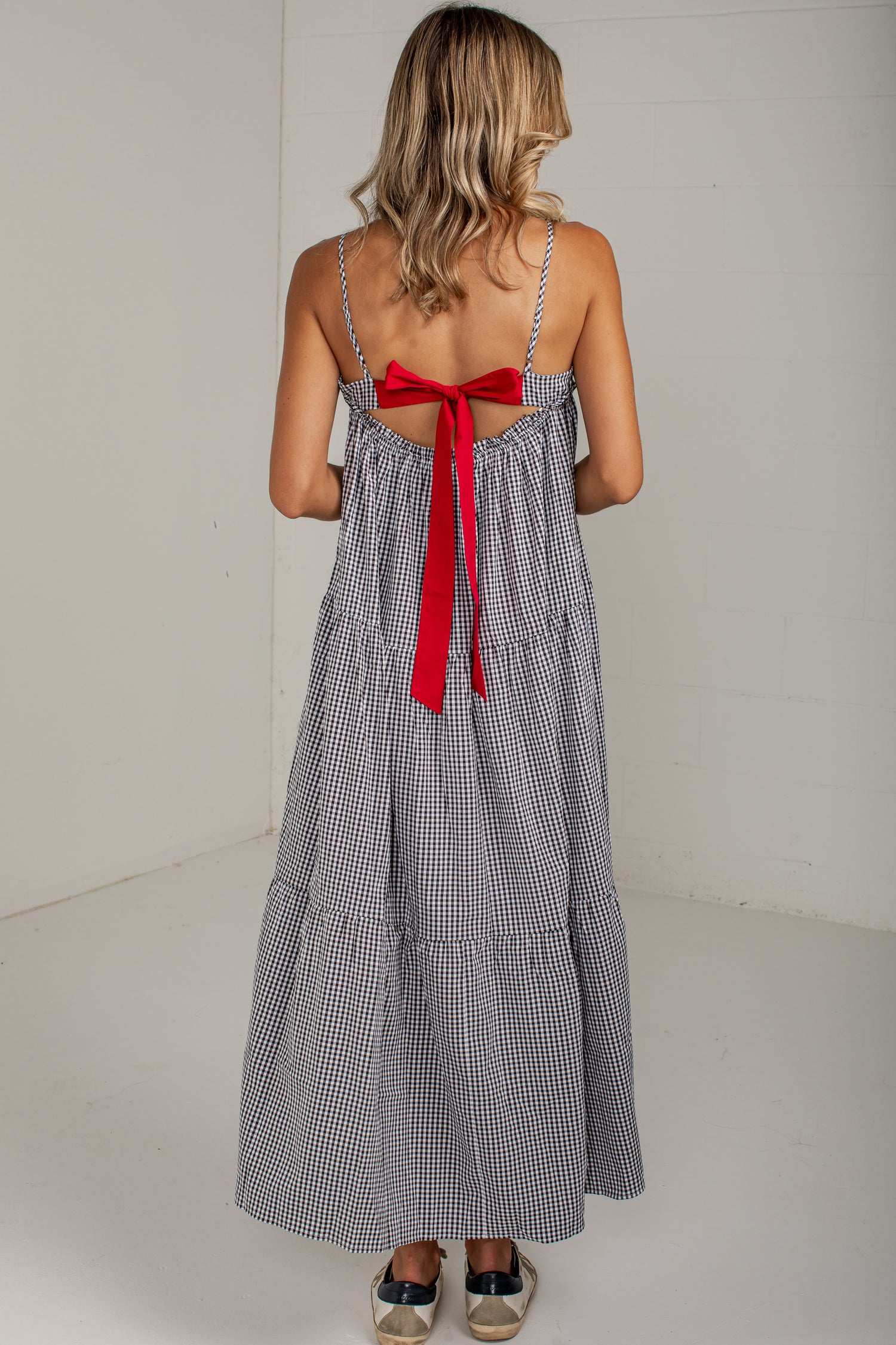 A woman viewed from behind wearing a gray and white gingham maxi dress with thin straps and a large red bow at the back, standing against a plain white wall.