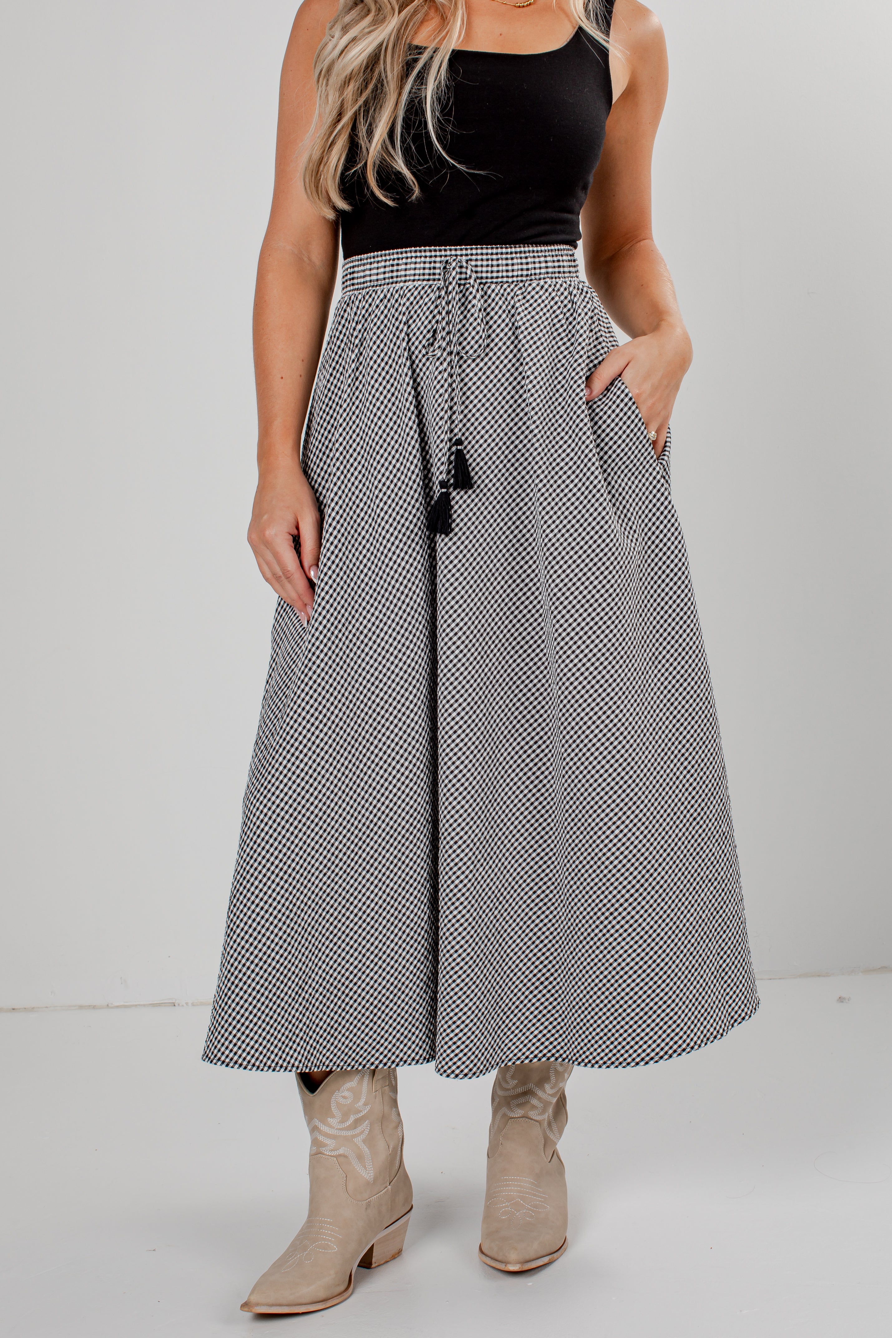A woman modeling a high-waisted, maxi-length skirt with a black and white micro-check pattern, a drawstring waist, and black tassels, paired with a black sleeveless top and beige boots.