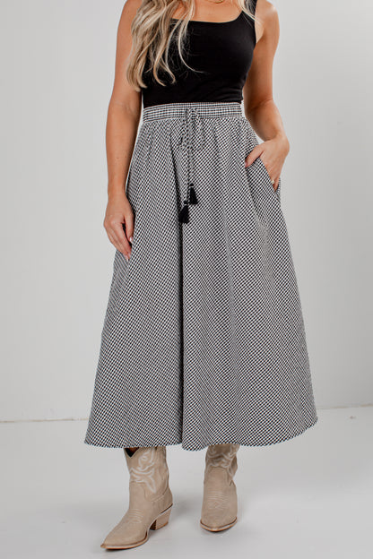 A woman modeling a high-waisted, maxi-length skirt with a black and white micro-check pattern, a drawstring waist, and black tassels, paired with a black sleeveless top and beige boots.
