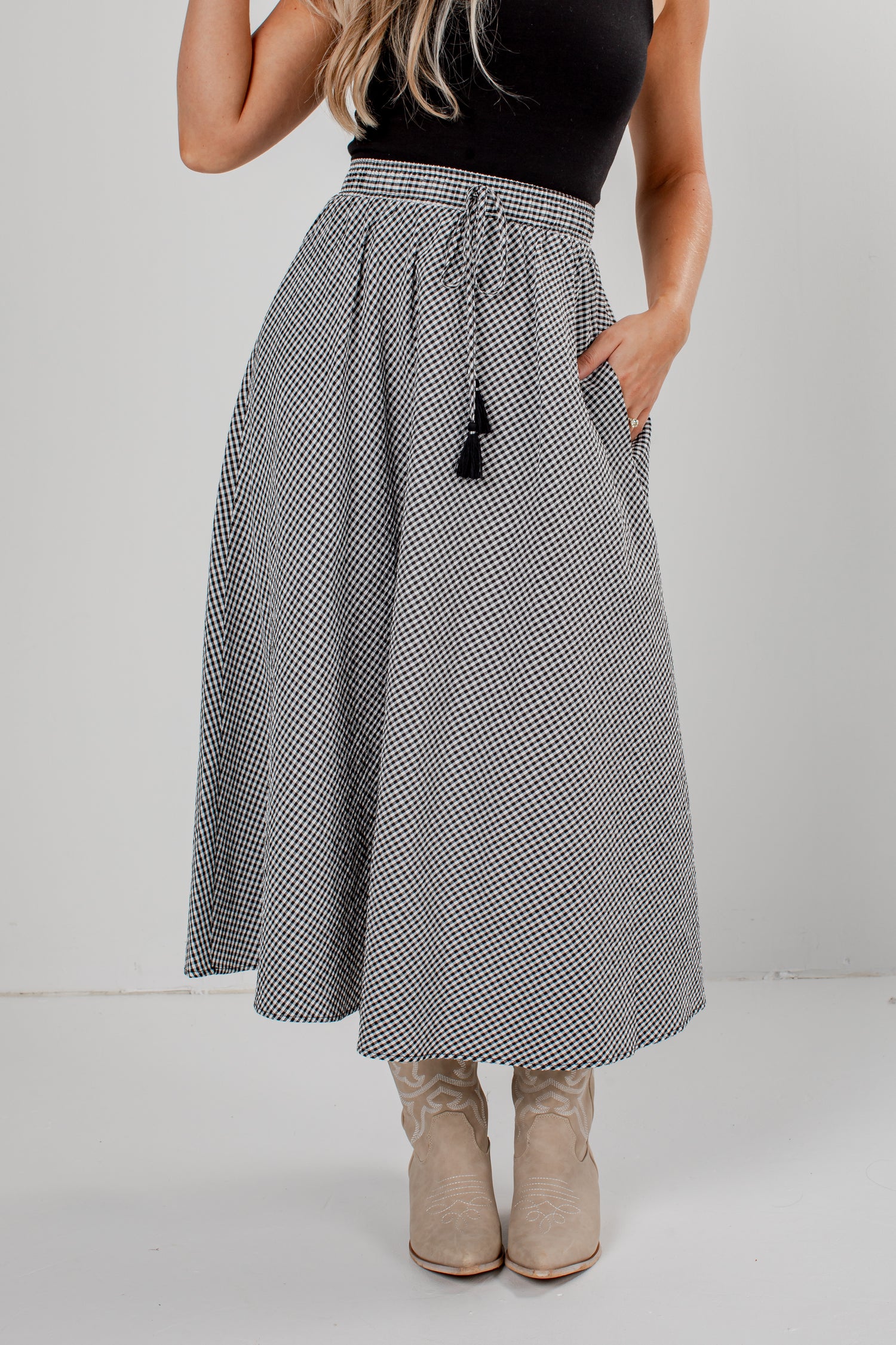 A woman wearing a high-waisted, A-line midi skirt with a black and white micro-pattern, a black top, and beige cowboy boots, standing against a light gray studio background.