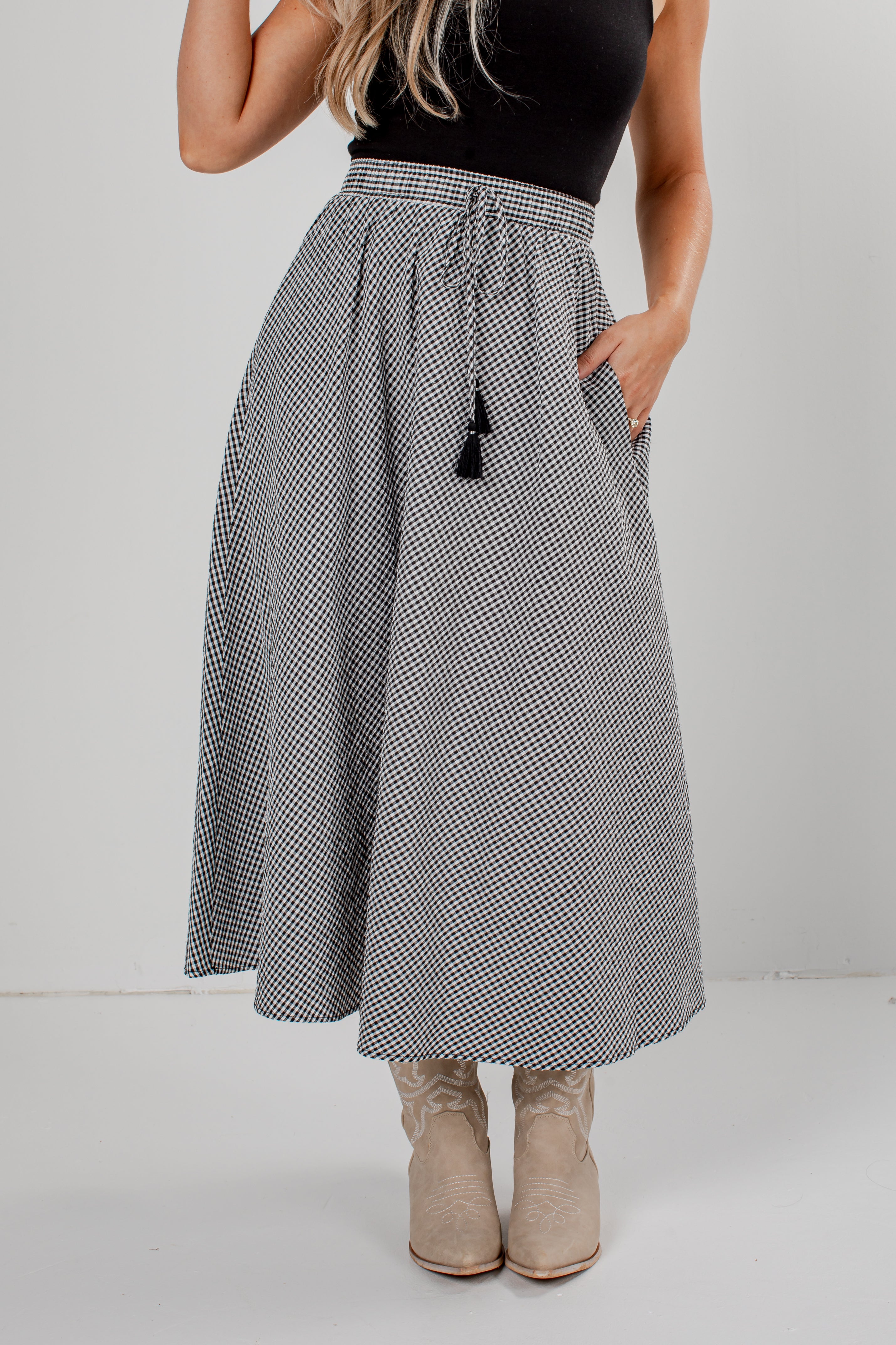 A woman wearing a high-waisted, A-line midi skirt with a black and white micro-pattern, a black top, and beige cowboy boots, standing against a light gray studio background.