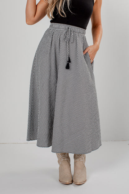 A woman wearing a high-waisted, A-line midi skirt with a black and white micro-pattern, a black top, and beige cowboy boots, standing against a light gray studio background.