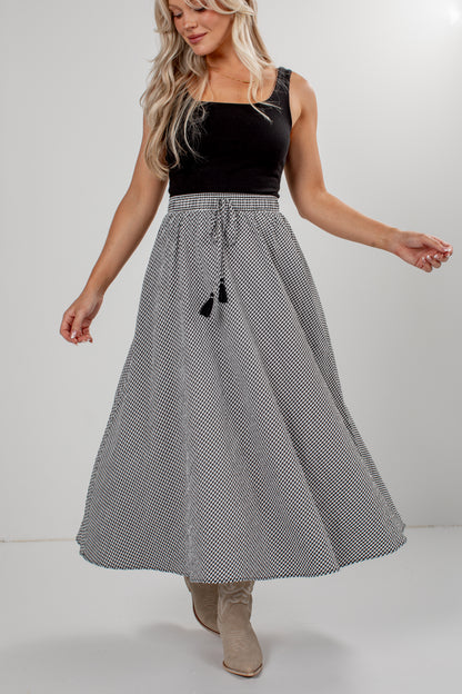 A woman modeling a high-waisted, knee-length skirt with a black and white micro-check pattern, a drawstring waist, and black tassels, paired with a black sleeveless top and beige boots.