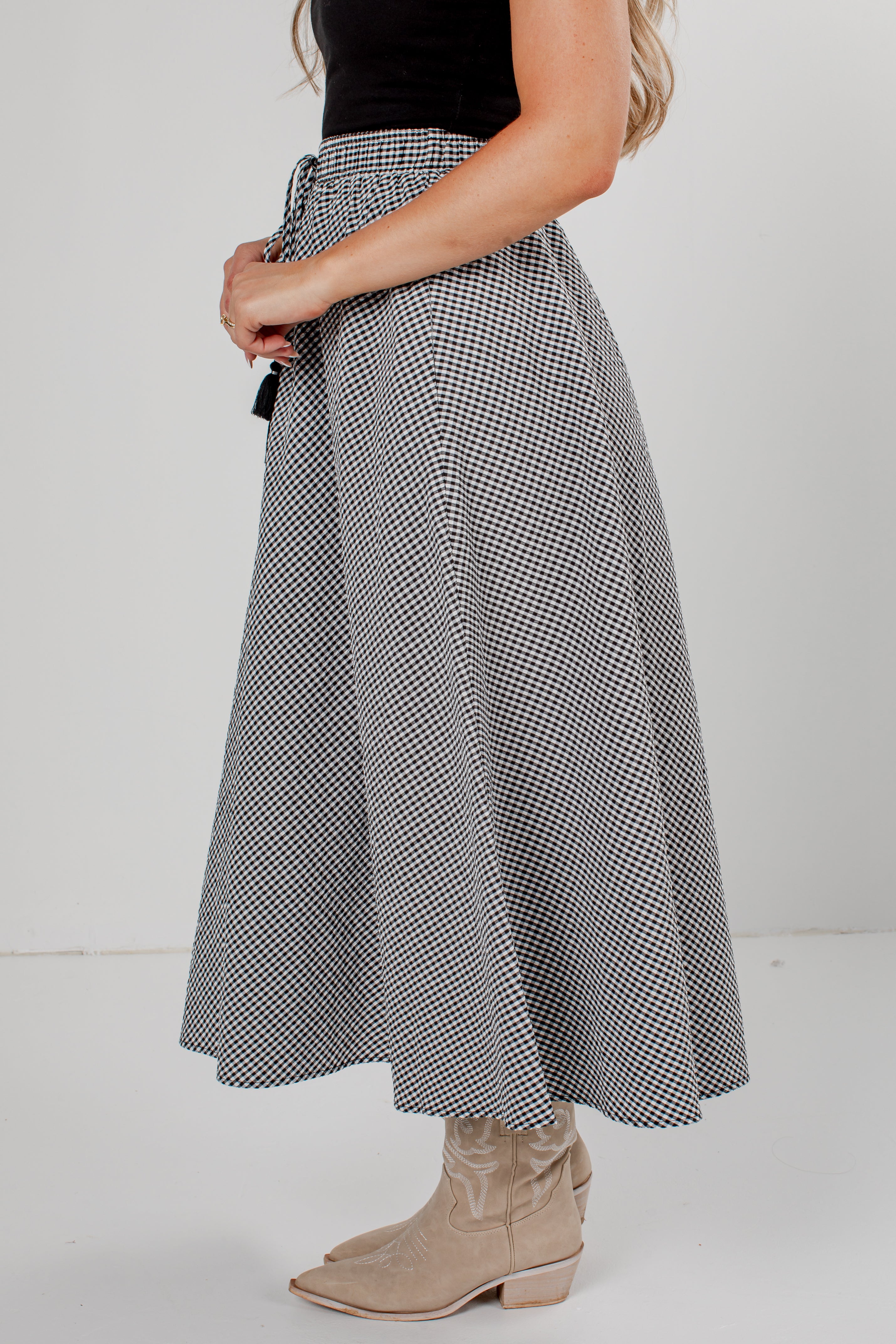 A woman wearing a high-waisted, A-line midi skirt with a black and white micro-check pattern, styled with a black top and beige cowboy boots against a plain white studio background.